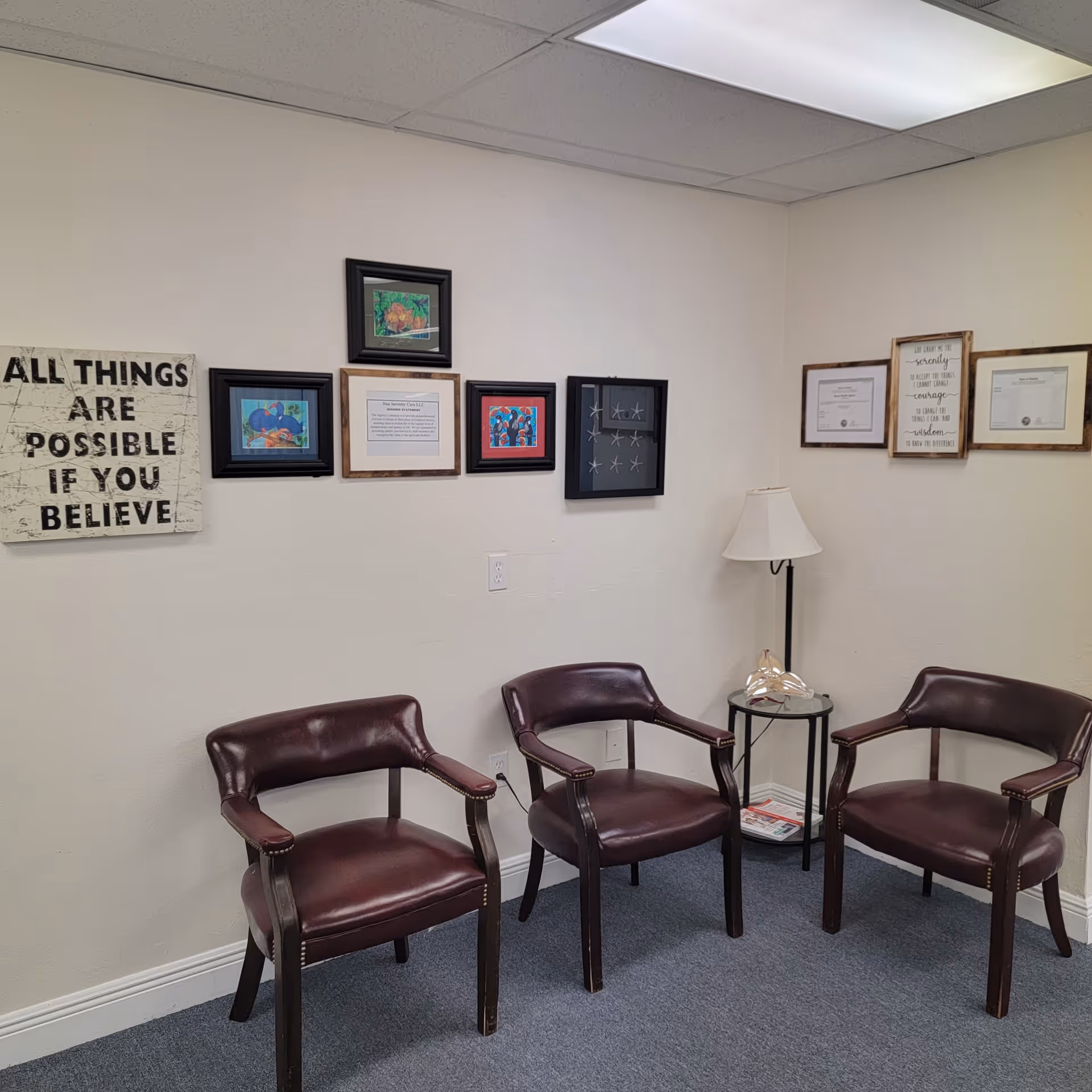 A small waiting area with three burgundy chairs, a glass side table, lamp, and framed art.