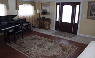 Sunlit living room with a grand piano, wheelchair, rug, and front entryway.