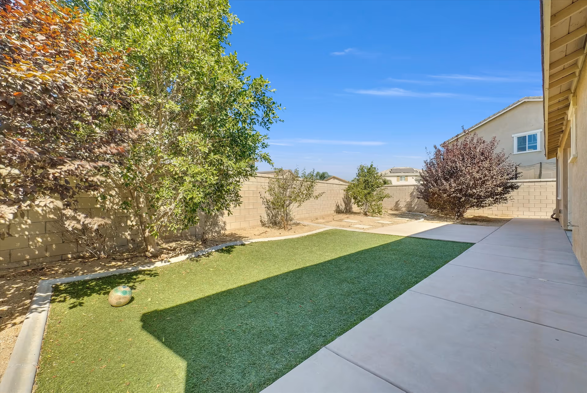 Sunny outdoor courtyard with artificial turf, trees, and a concrete patio.