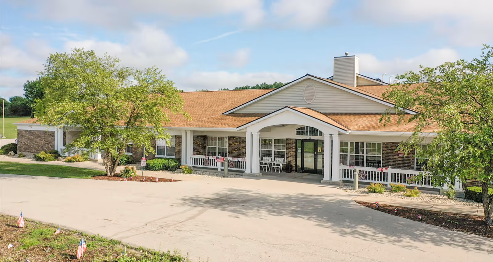 Single-story assisted living building with a covered entrance, porch seating, and surrounding trees.