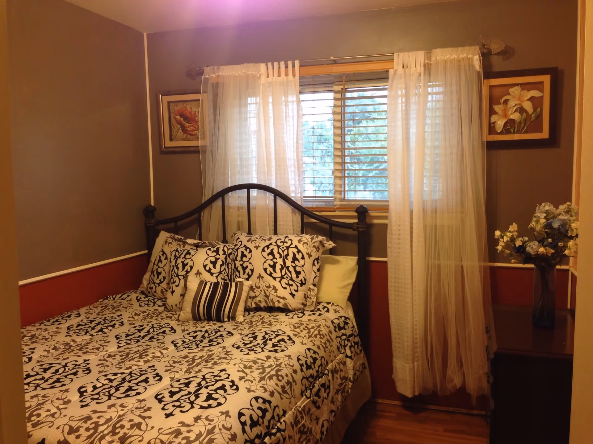 A cozy bedroom with a bed featuring a black and white patterned comforter and pillows. The bed has a black metal headboard and is positioned against a wall with a window above it, covered by sheer white curtains and blinds. On either side of the window are framed floral paintings. To the right of the bed is a small wooden nightstand with a vase of flowers.