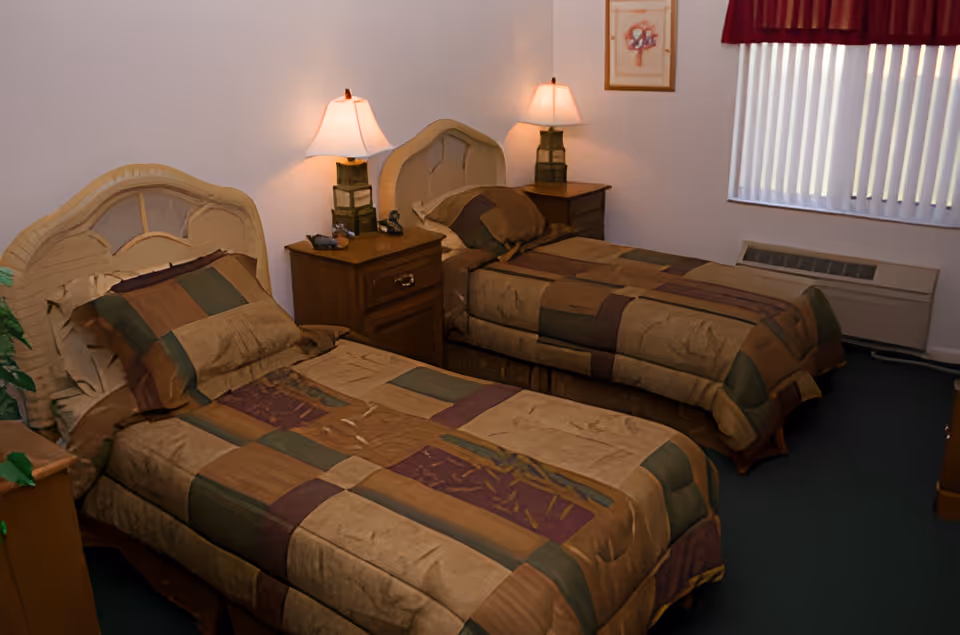 A bedroom with two single beds featuring matching brown, green, and purple patterned bedspreads. Each bed has a wooden headboard and a pillow. Between the beds is a wooden nightstand with two lamps and a telephone. A window with vertical blinds and a red valance is on the right wall, and a framed picture hangs above the nightstand.
