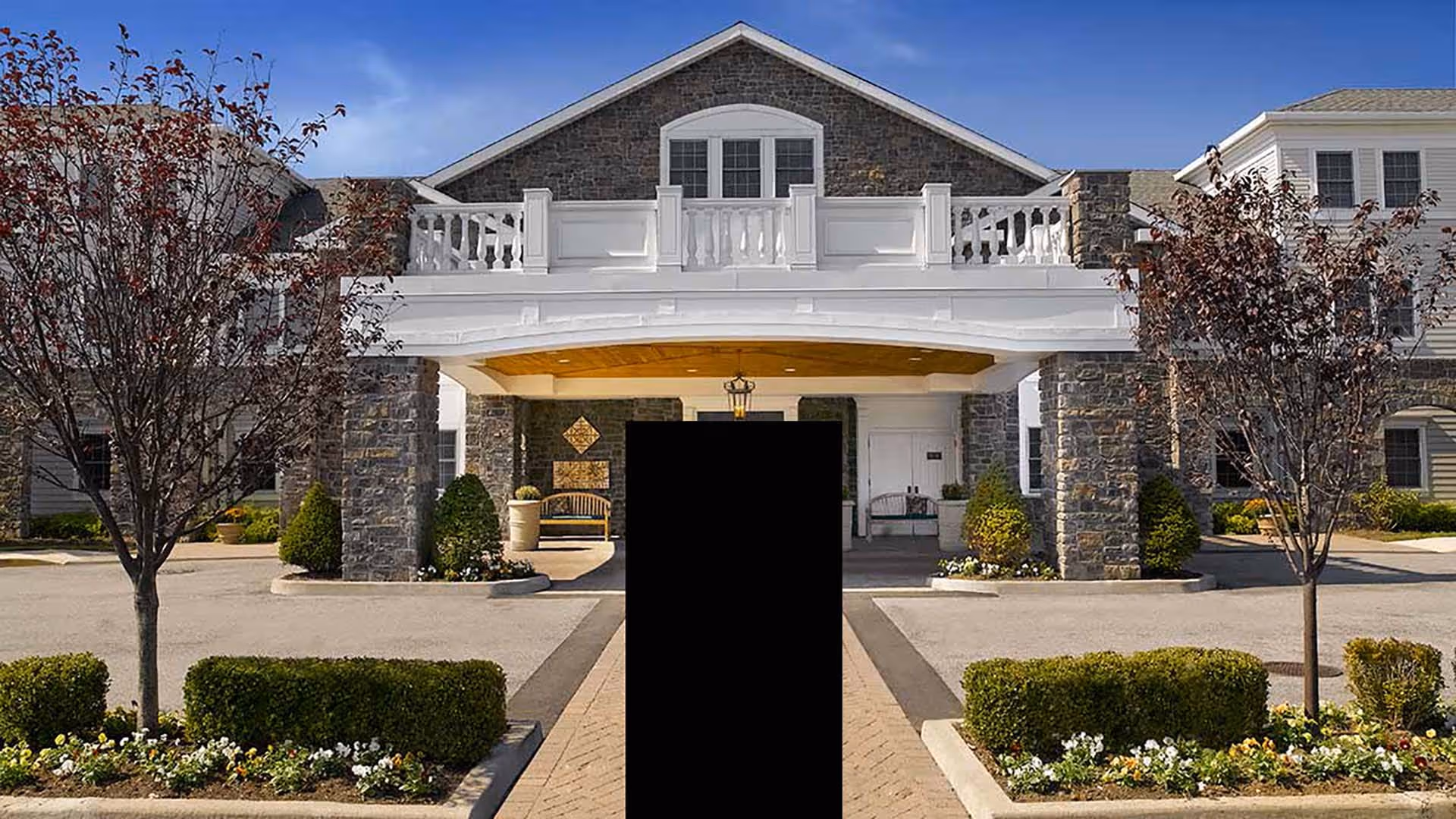 Front exterior view of a senior living facility with stone pillars, white balcony railing, and a covered entrance. There are landscaped bushes, flowers, and trees on either side of the driveway leading to the entrance.