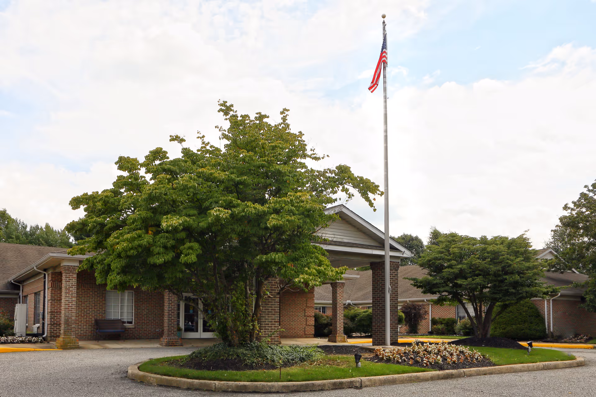 Exterior view of a single-story brick building with a covered entrance, surrounded by trees and landscaping. An American flag flies on a tall flagpole near the entrance under a partly cloudy sky.