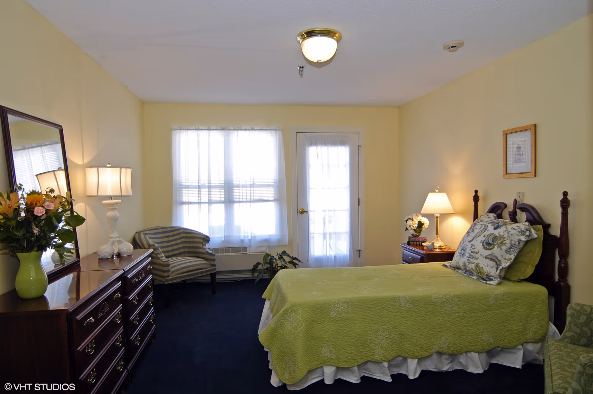 A cozy bedroom with a single bed covered in a green quilt and floral pillows. There is a wooden dresser with a large mirror and a lamp on top, along with a green vase holding flowers. A striped armchair is positioned near a window with sheer white curtains. A nightstand with a lamp and a small flower arrangement is next to the bed. The walls are painted light yellow and the carpet is dark blue.