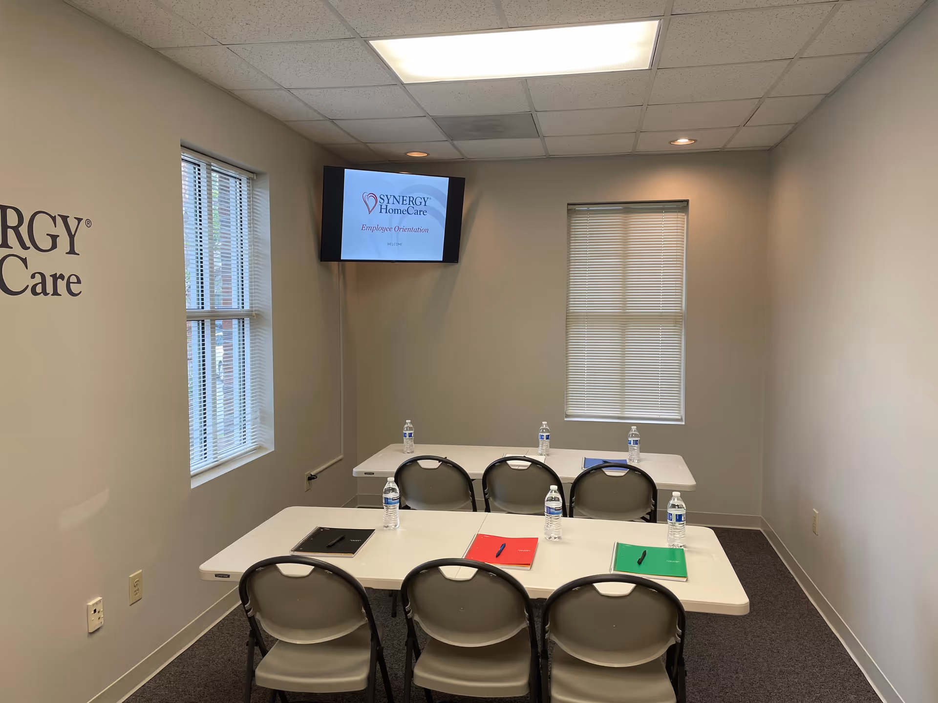 Small training room with tables, chairs, water bottles, notebooks, and a SYNERGY HomeCare screen.