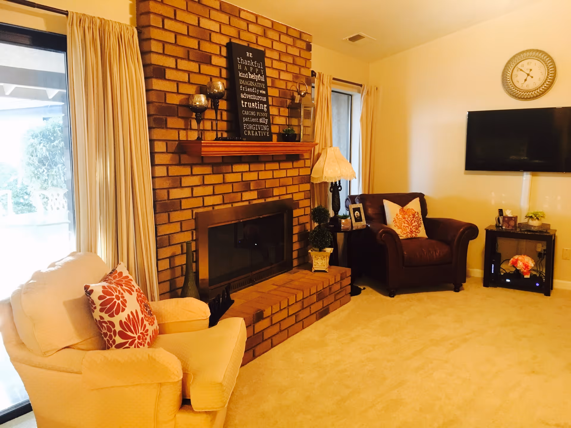 A cozy living room with a brick fireplace in the center. There is a cream-colored armchair with a red floral pillow on the left and a dark brown leather armchair with a similar pillow on the right. The fireplace mantel holds decorative items including candle holders and a framed inspirational sign. Beige curtains frame two windows, and a flat-screen TV is mounted on the wall above a small black table with flowers and other decor. A round wall clock hangs above the TV.