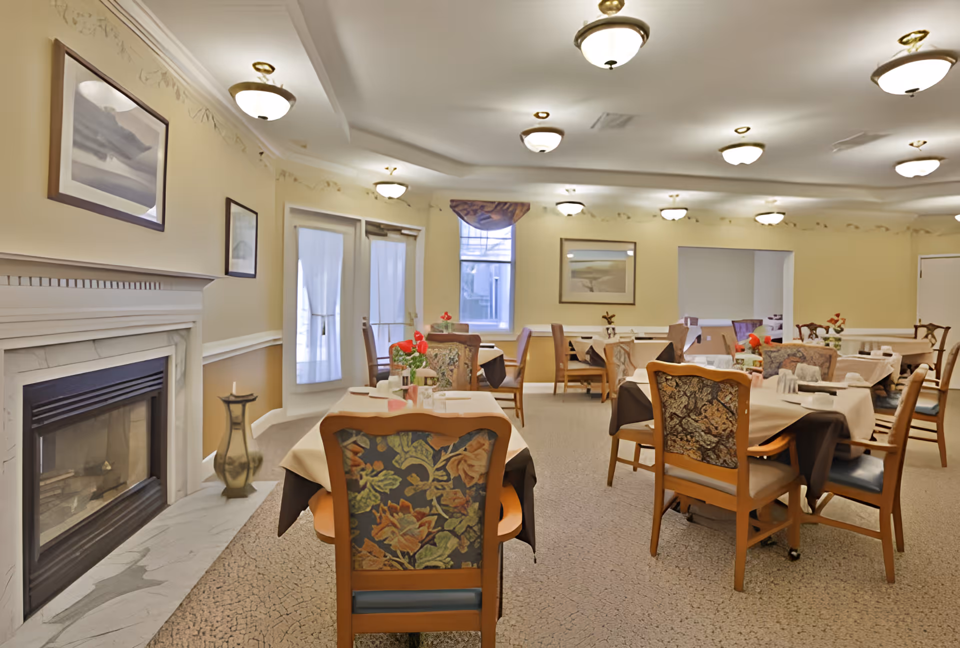 A dining room with multiple tables covered with beige and brown tablecloths, each set with napkins and small flower arrangements. The room features floral upholstered wooden chairs, a fireplace on the left wall, framed artwork, and several ceiling lights. There are windows and glass doors with curtains letting in natural light.