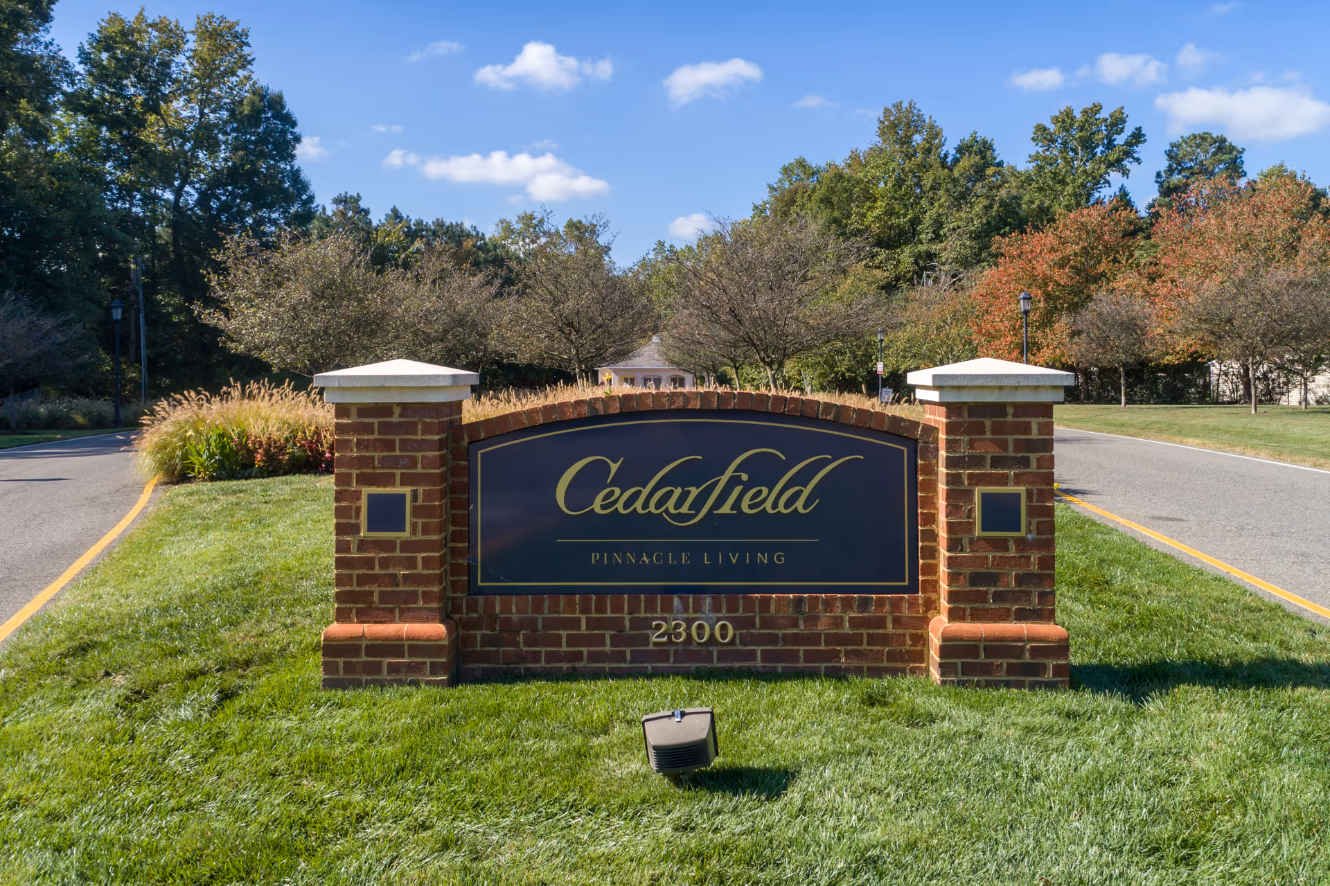 Brick entrance sign reading "Cedarfield Pinnacle Living" on a grassy median at the facility driveway.