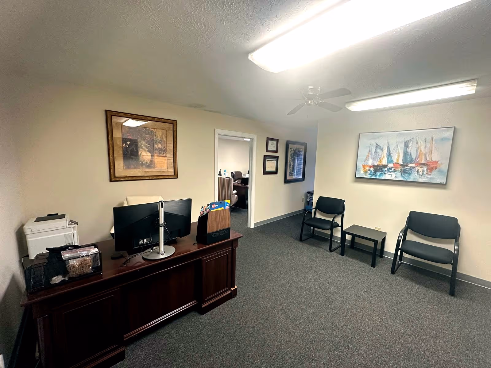 Office reception area with desk, computer, chairs, and wall art in a beige room.