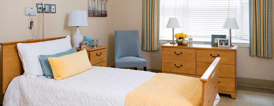 A neatly made single bed with white and yellow bedding in a senior living facility room. Next to the bed is a wooden nightstand with a white lamp, framed photos, and decorative items. A blue upholstered chair is placed near a window with striped curtains. Across from the bed is a wooden dresser with two white lamps, a vase of yellow flowers, and framed photos. The room has a calm and cozy atmosphere with soft natural light coming through the window blinds.