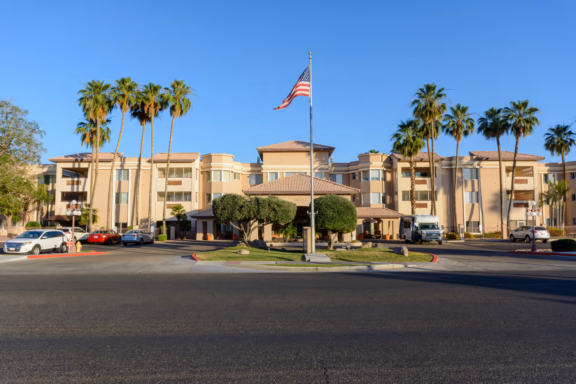 Front exterior view of The Palms at Sun City Assisted & Senior Living facility with a clear blue sky, palm trees, an American flag on a flagpole in the center, and several parked cars in front of the building.