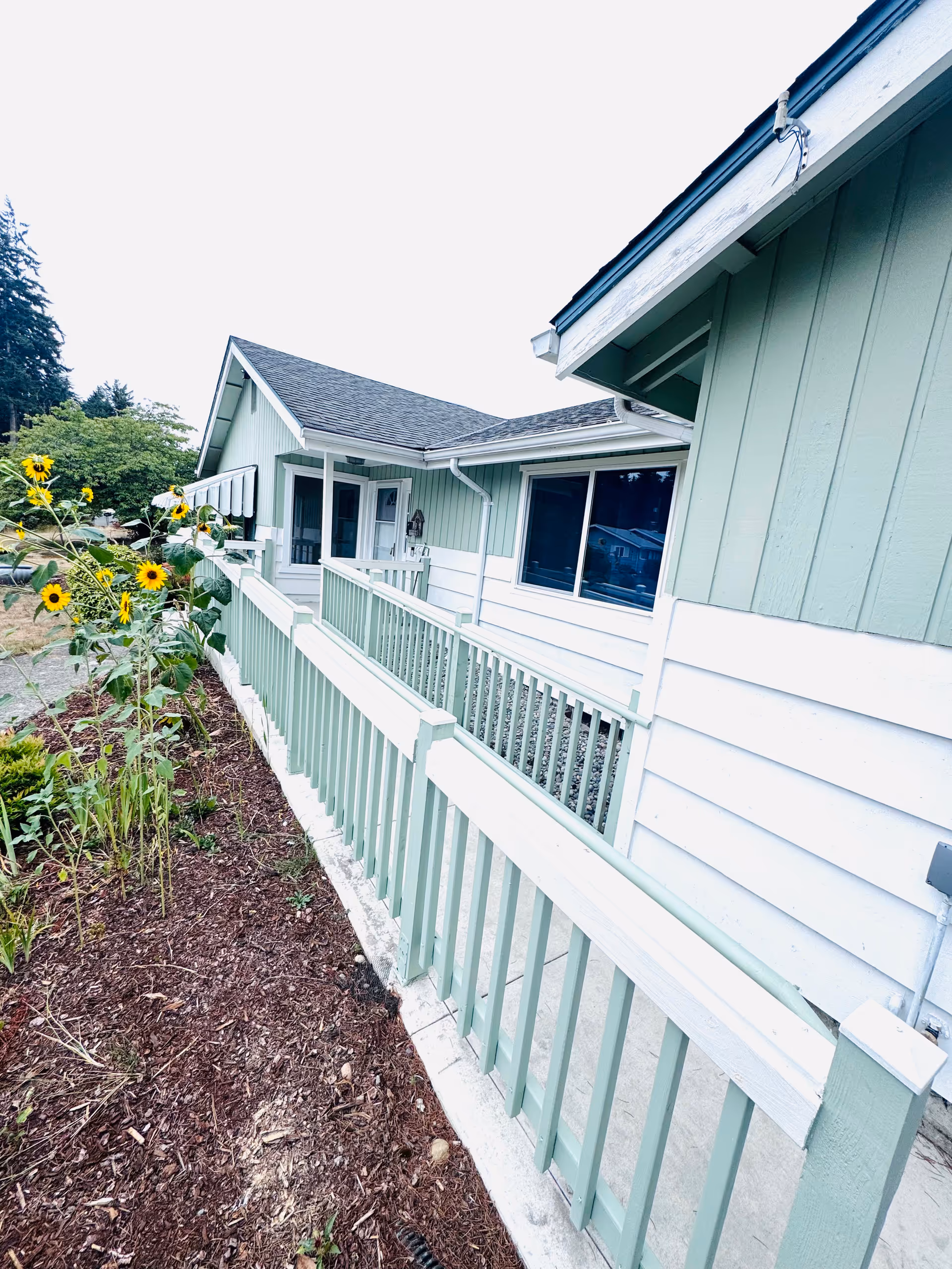 Green exterior of a senior home with a railing, walkway, and sunflowers in a garden bed