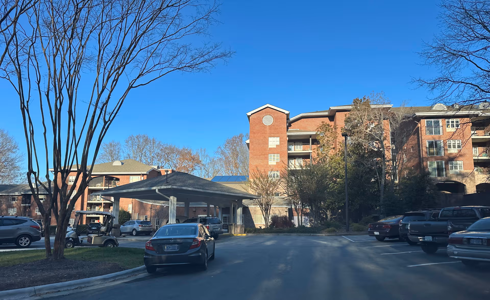 Bright blue sky over a brick senior living building with a covered entrance and parked cars.