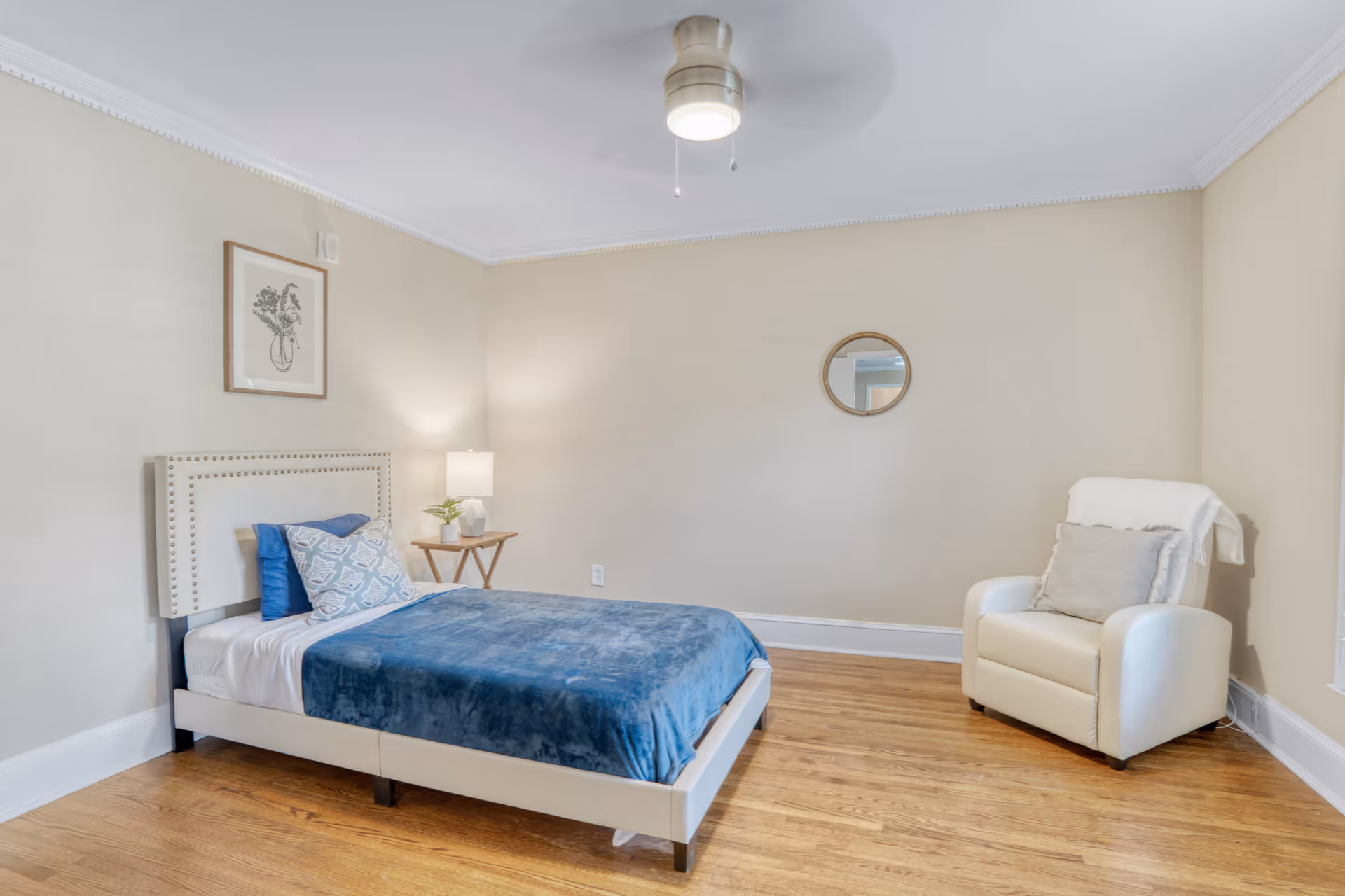 Neutral-toned bedroom with a bed, chair, nightstand, and round wall mirror.