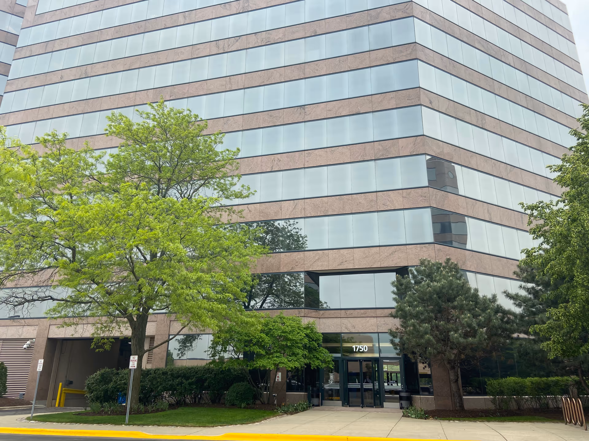 A tall glass-and-brick office building with trees in front and a visible entrance.
