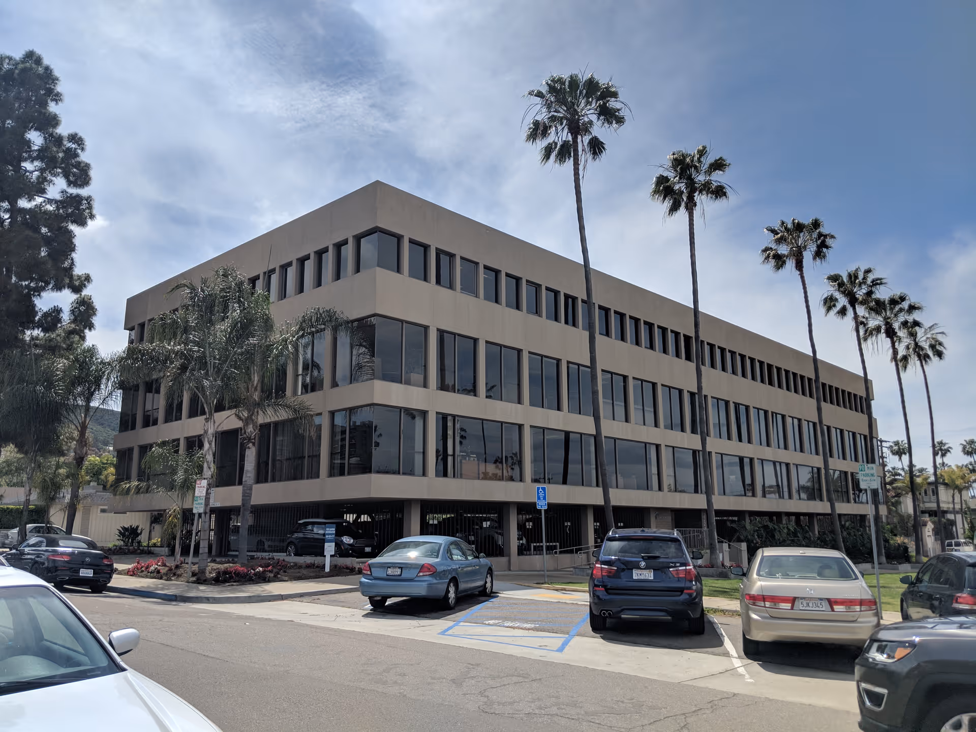 Three-story beige building with large windows, palm trees, and parked cars.