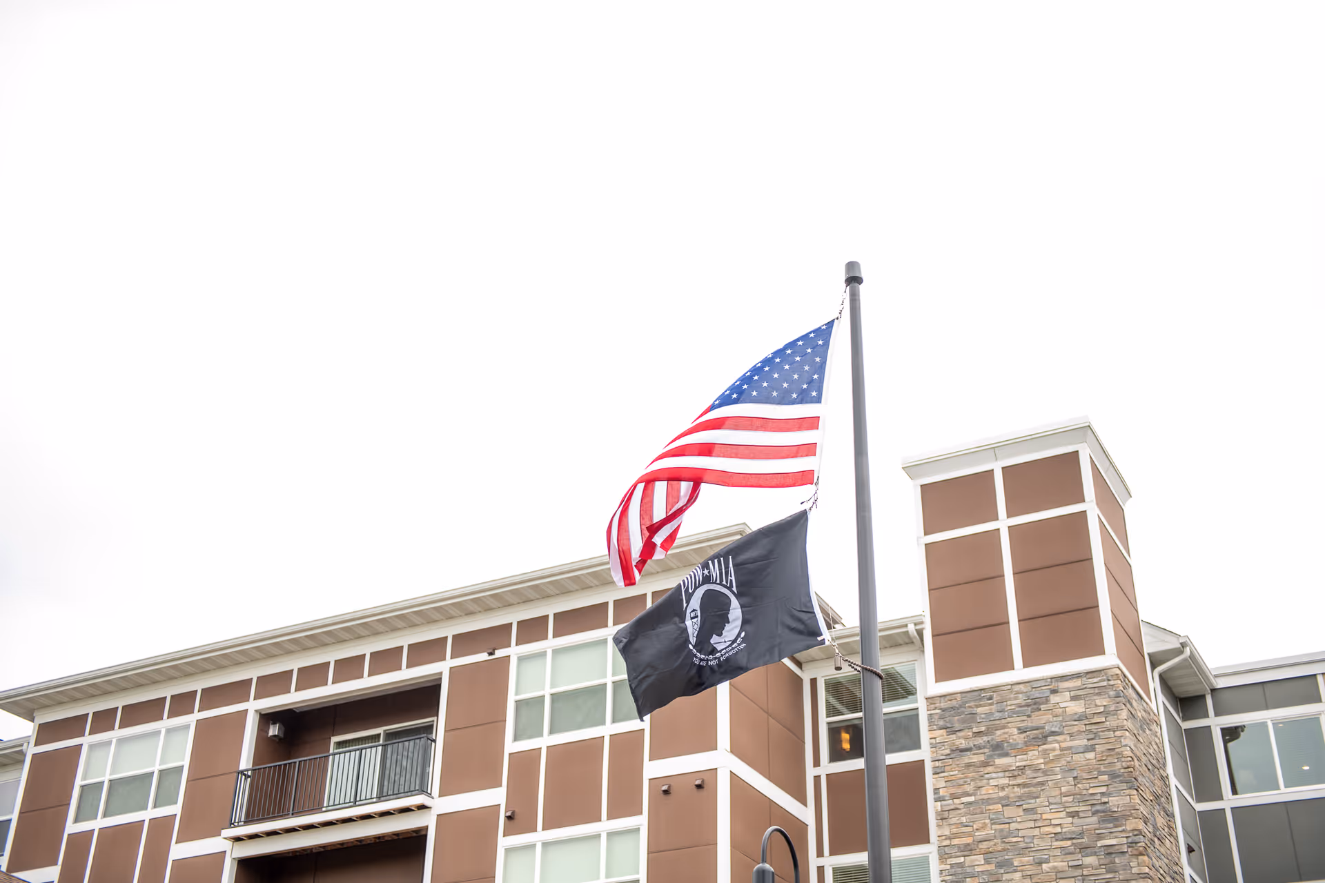 Exterior view of a multi-story senior living facility building with brown and white paneling and stone accents. Two flags are flying on a flagpole in front of the building: the American flag and a black POW/MIA flag.