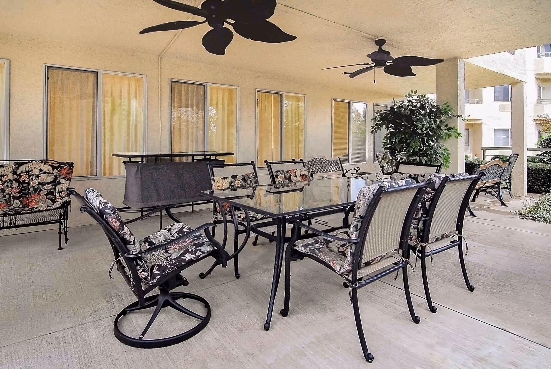 Covered outdoor patio area with a glass-top table surrounded by six cushioned chairs with floral patterns. There are two ceiling fans above, a bench, a potted plant, and windows with closed curtains on the wall behind the seating area.