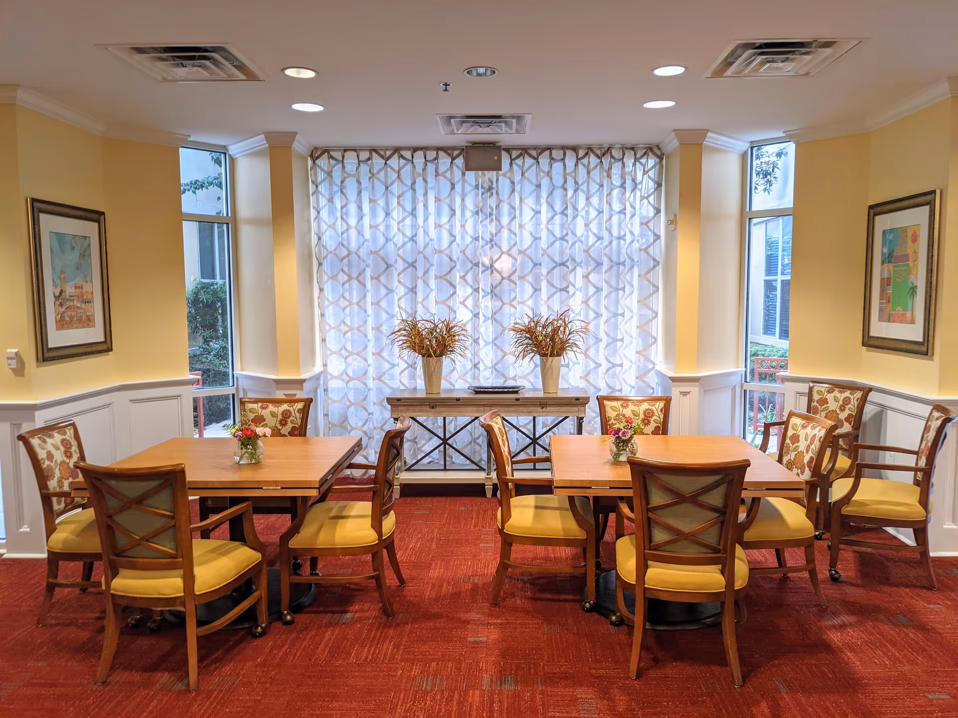 A cozy dining area with two wooden tables, each surrounded by four chairs with yellow cushions and floral-patterned backs. The room has yellow walls with white wainscoting, red carpet flooring, and two framed pictures on the walls. A console table with two vases of plants is positioned in front of a large window covered with patterned sheer curtains.