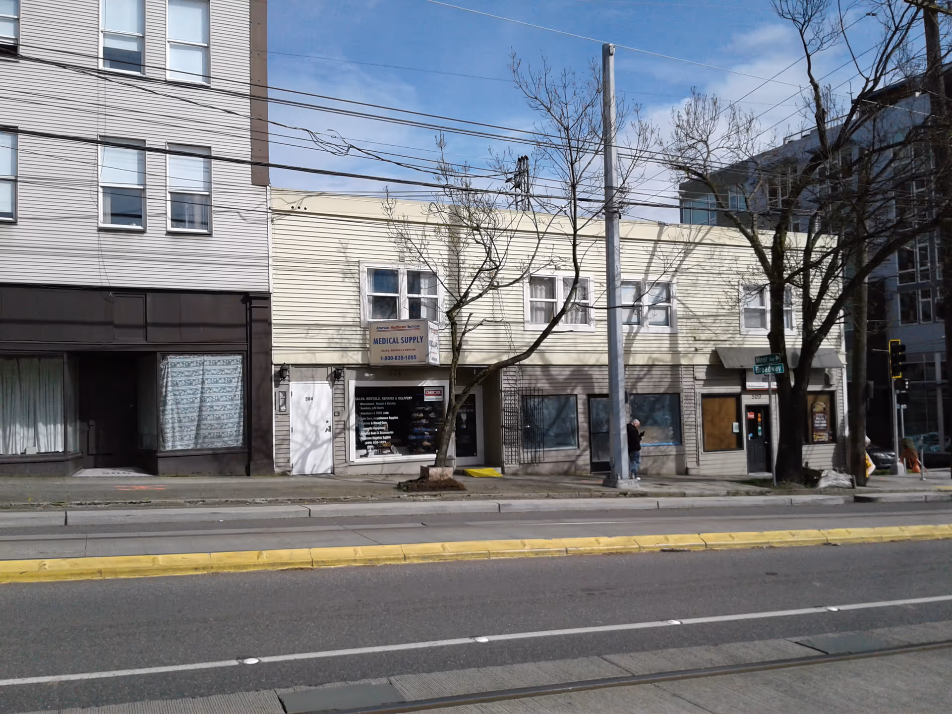 Street view of storefronts along a sidewalk with leafless trees and power lines.