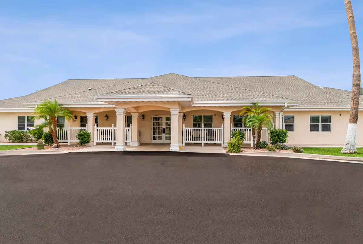 Front exterior view of a single-story senior living facility building with a covered entrance supported by columns, surrounded by small palm trees and landscaping under a clear blue sky.