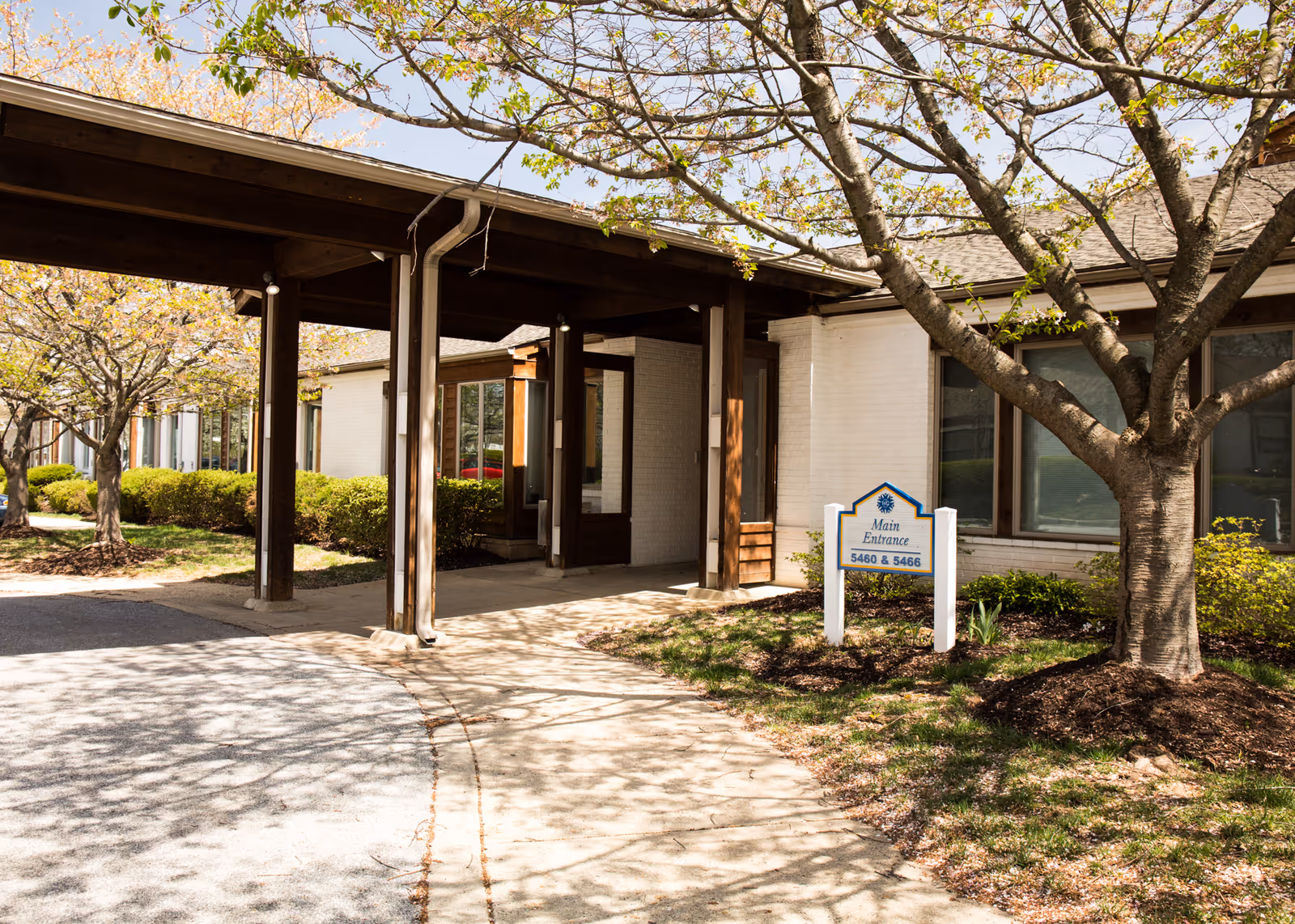 Covered main entrance to a single-story senior living building with a sidewalk, trees, and a 'Main Entrance' sign.