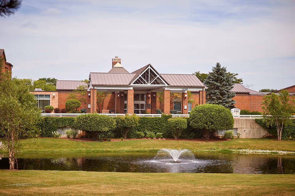 Brick senior living building with a covered entrance, landscaped shrubs and a pond with a fountain in front.