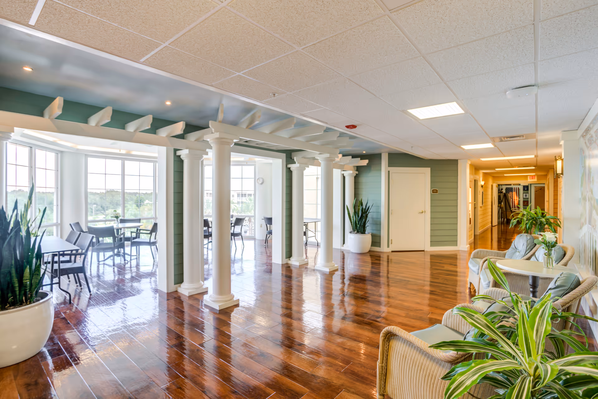 Bright senior living lounge with white columns, wooden floors, and large windows.