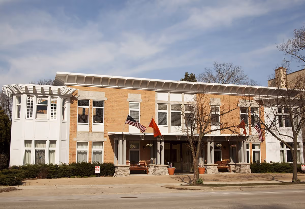 Two-story brick senior living facility facade with flags, windows, benches, and a sidewalk entrance.