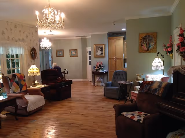 Cozy senior living room with wooden floors, lamps, and residents seated.