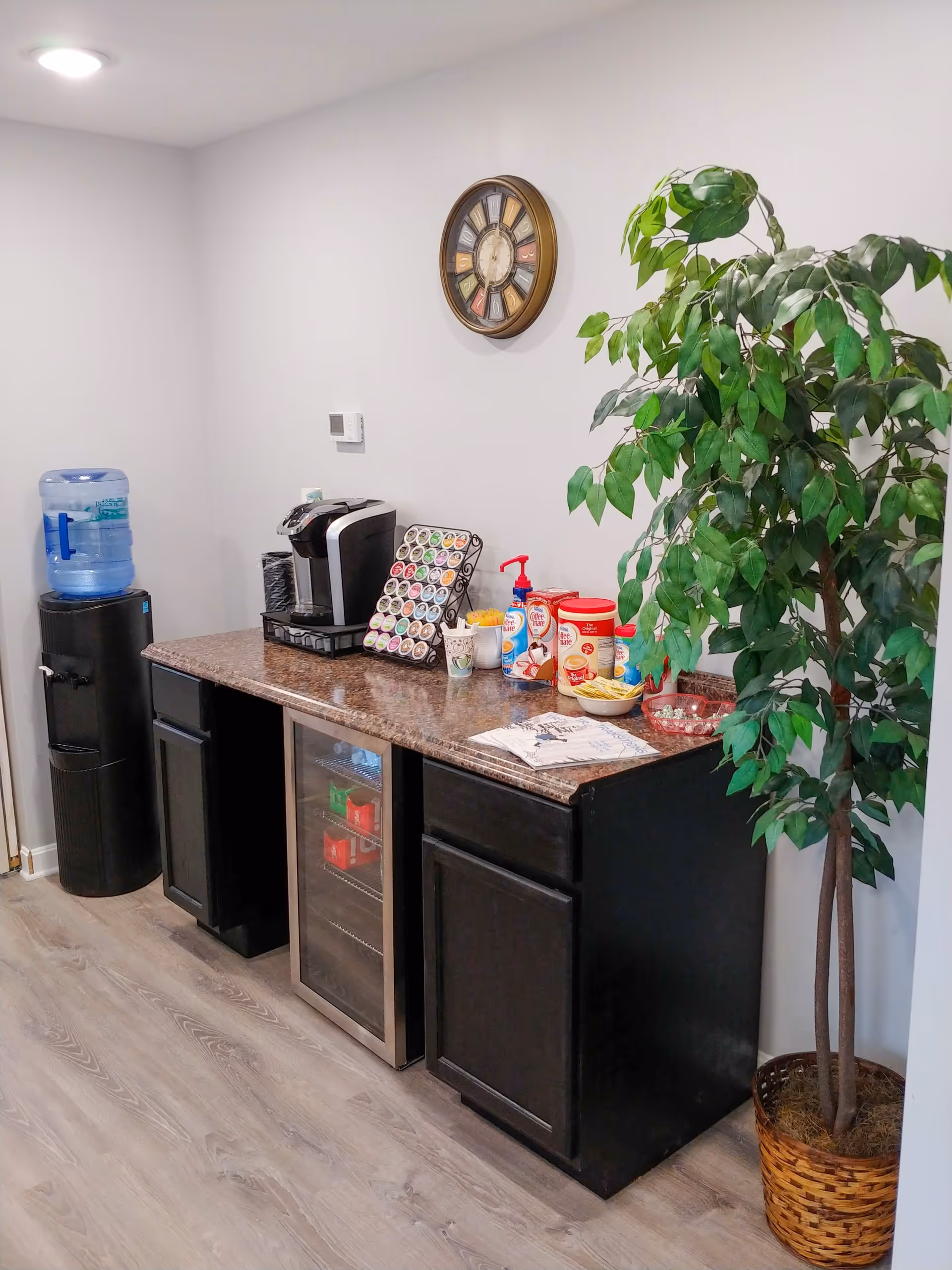 Small kitchenette area with coffee station, mini-fridge, water cooler, snacks, and a potted plant.