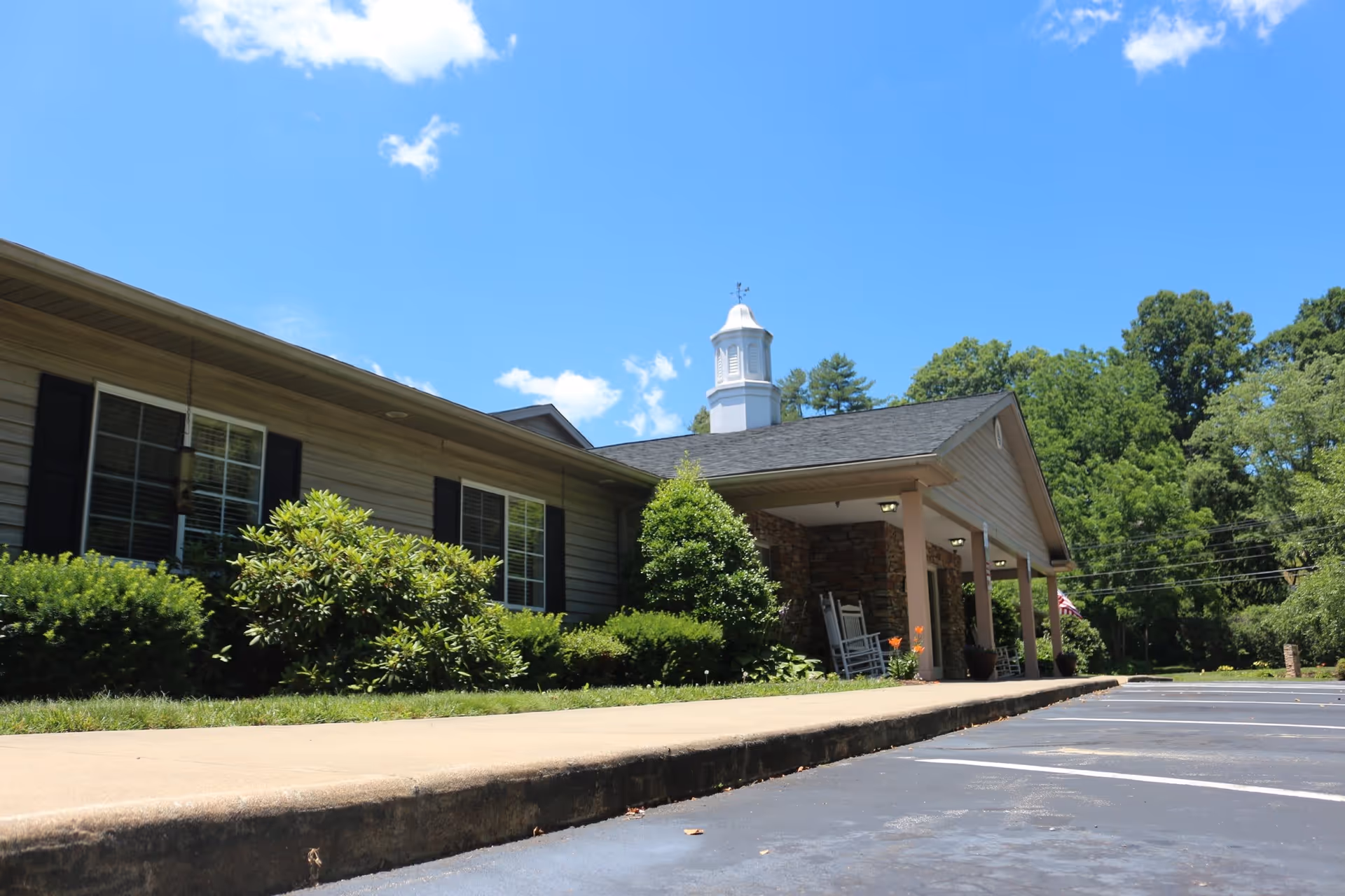 Exterior view of Kingsbridge House, a single-story building with beige siding, black window shutters, and a small white cupola on the roof. The building is surrounded by green bushes and trees under a clear blue sky. There is a sidewalk and an empty parking lot in the foreground.