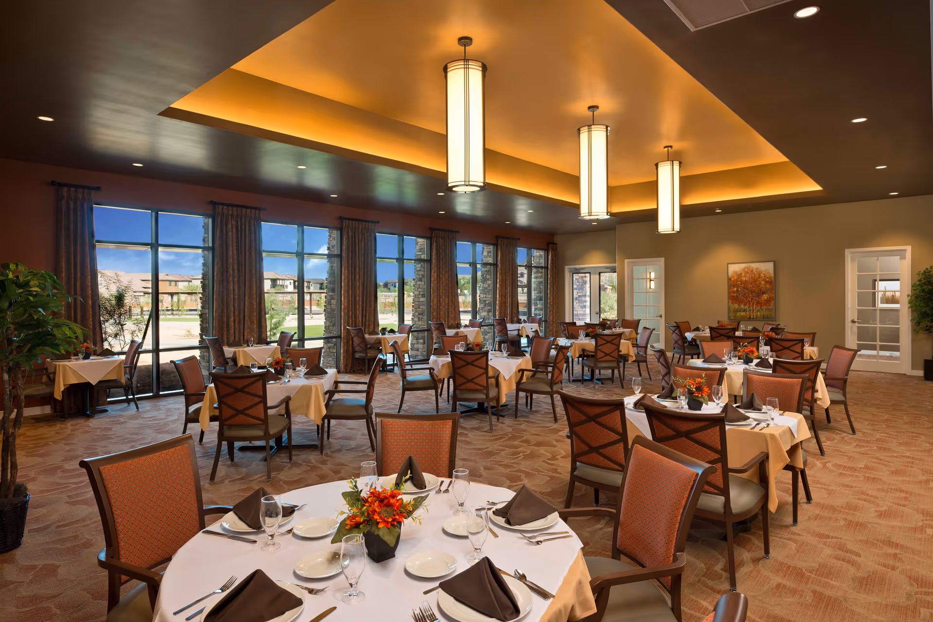 A spacious dining room with multiple tables covered in white and beige tablecloths, each set with plates, glasses, silverware, and brown folded napkins. The room features large windows with views of an outdoor area and buildings, warm ceiling lighting, and decorative plants and artwork on the walls.