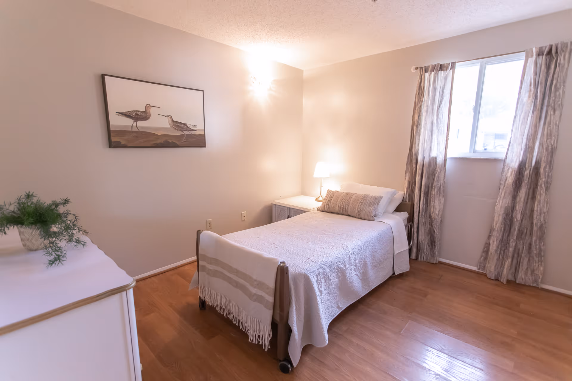 A tidy bedroom in a senior living facility with a bed, dresser, lamp, and window.