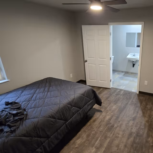 Bedroom with dark comforter bed, ceiling fan, and open door revealing a bathroom sink.