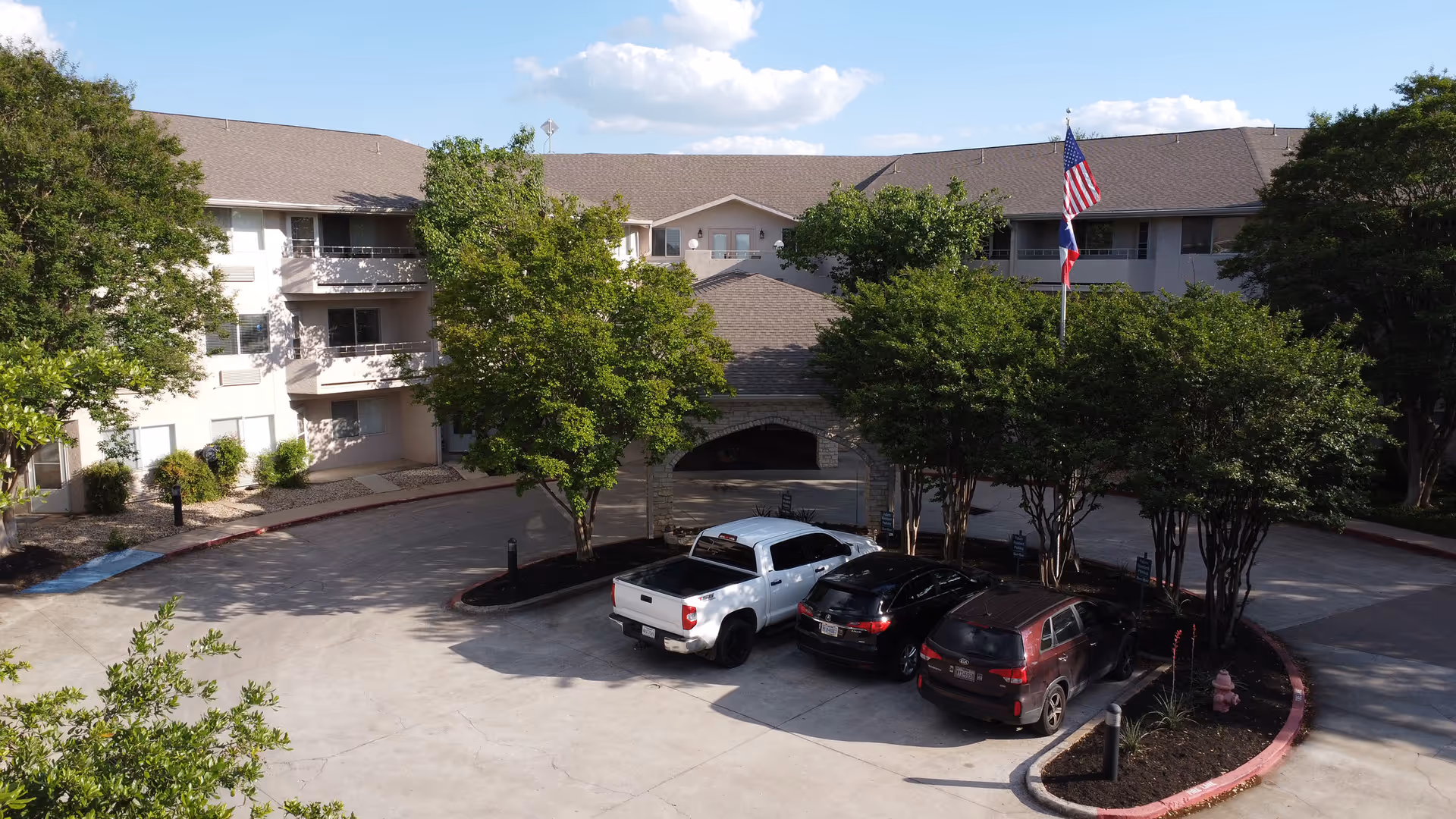 Exterior view of Solstice Senior Living at Austin showing a three-story building with balconies, surrounded by trees and a parking area with three vehicles. An American flag and a Texas state flag are flying on a flagpole near the entrance.