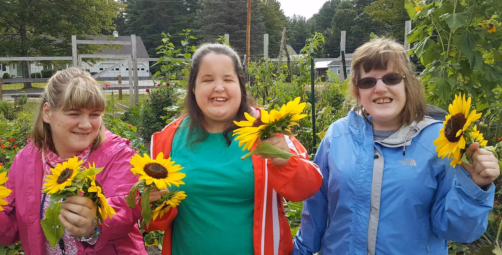 Three smiling women hold sunflowers in a sunny outdoor garden.