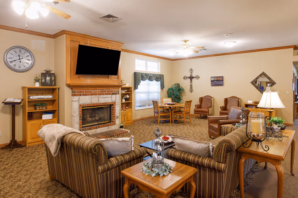 A cozy living room area in an assisted living facility featuring a stone and brick fireplace with a mounted flat-screen TV above it. The room includes striped sofas with pillows and a throw blanket, wooden side tables with decorative items, a round wooden table with chairs near a window with green valances, and several brown leather armchairs. The walls are decorated with a large clock, a cross, a mirror, and framed artwork. The room is warmly lit with ceiling fans and lamps.