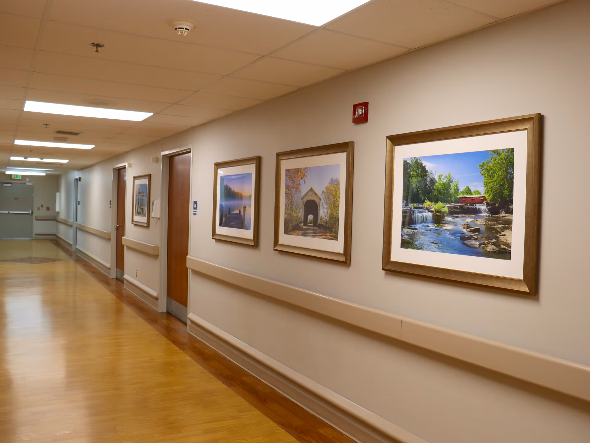 A clean, well-lit hallway in a senior living facility with wooden doors on the left side and framed landscape photographs on the right wall. The floor is wooden with beige walls and handrails along the corridor.