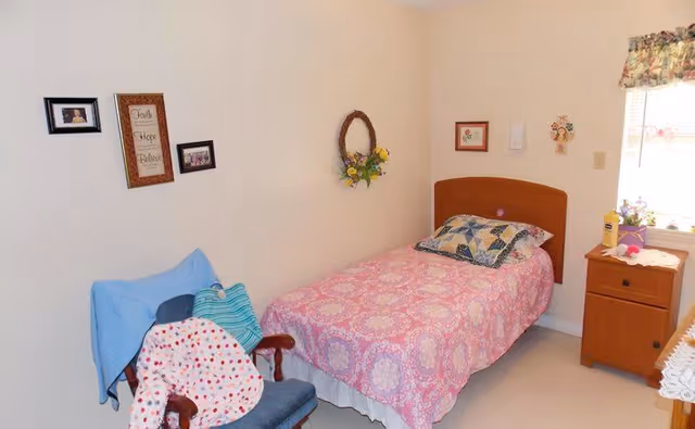 Small bedroom with a single bed in pink bedding, a wooden nightstand by a window, a chair with blankets, and wall decorations.