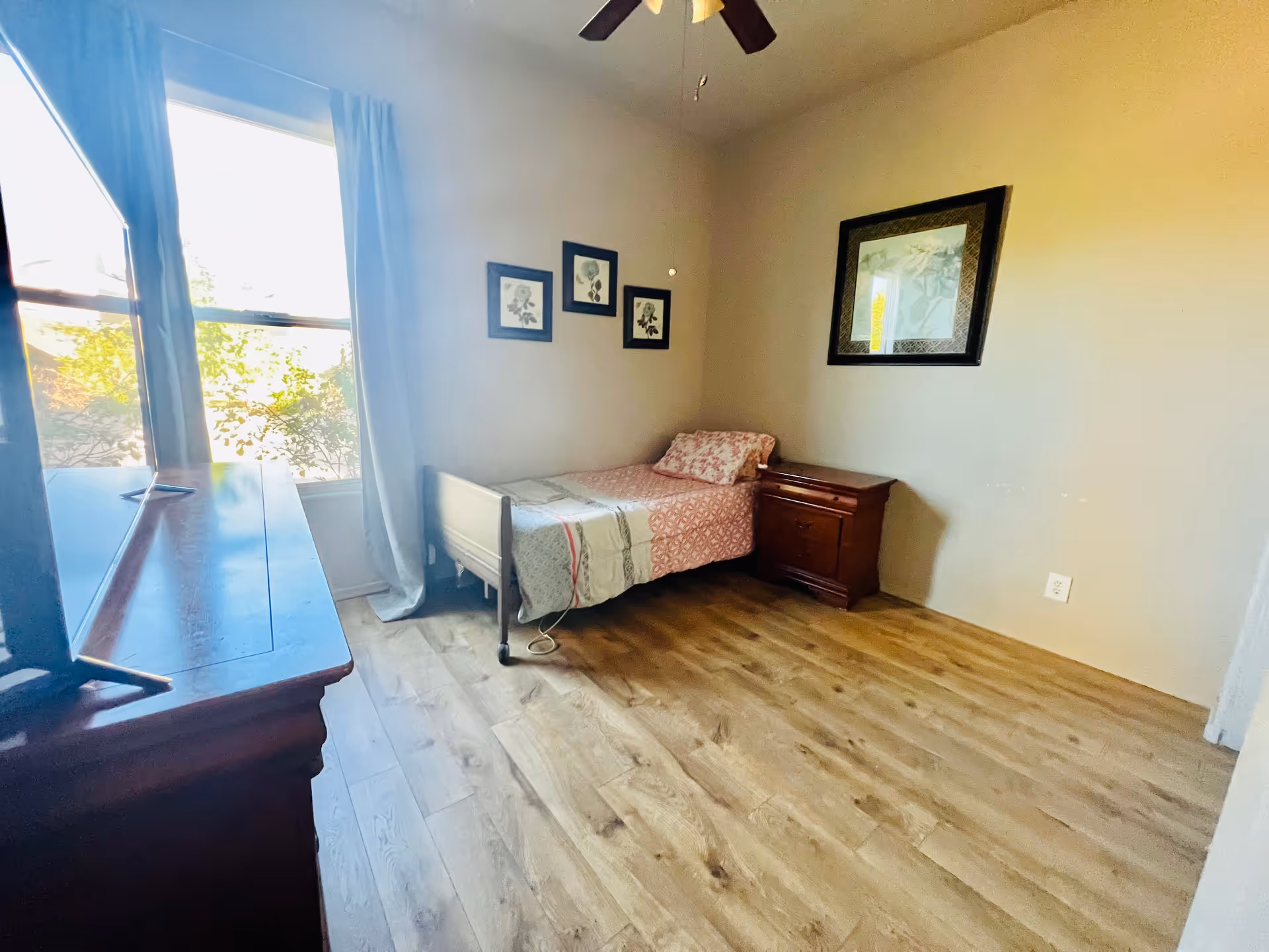 Sunlit bedroom with a single bed, nightstand, dresser, and framed artwork.