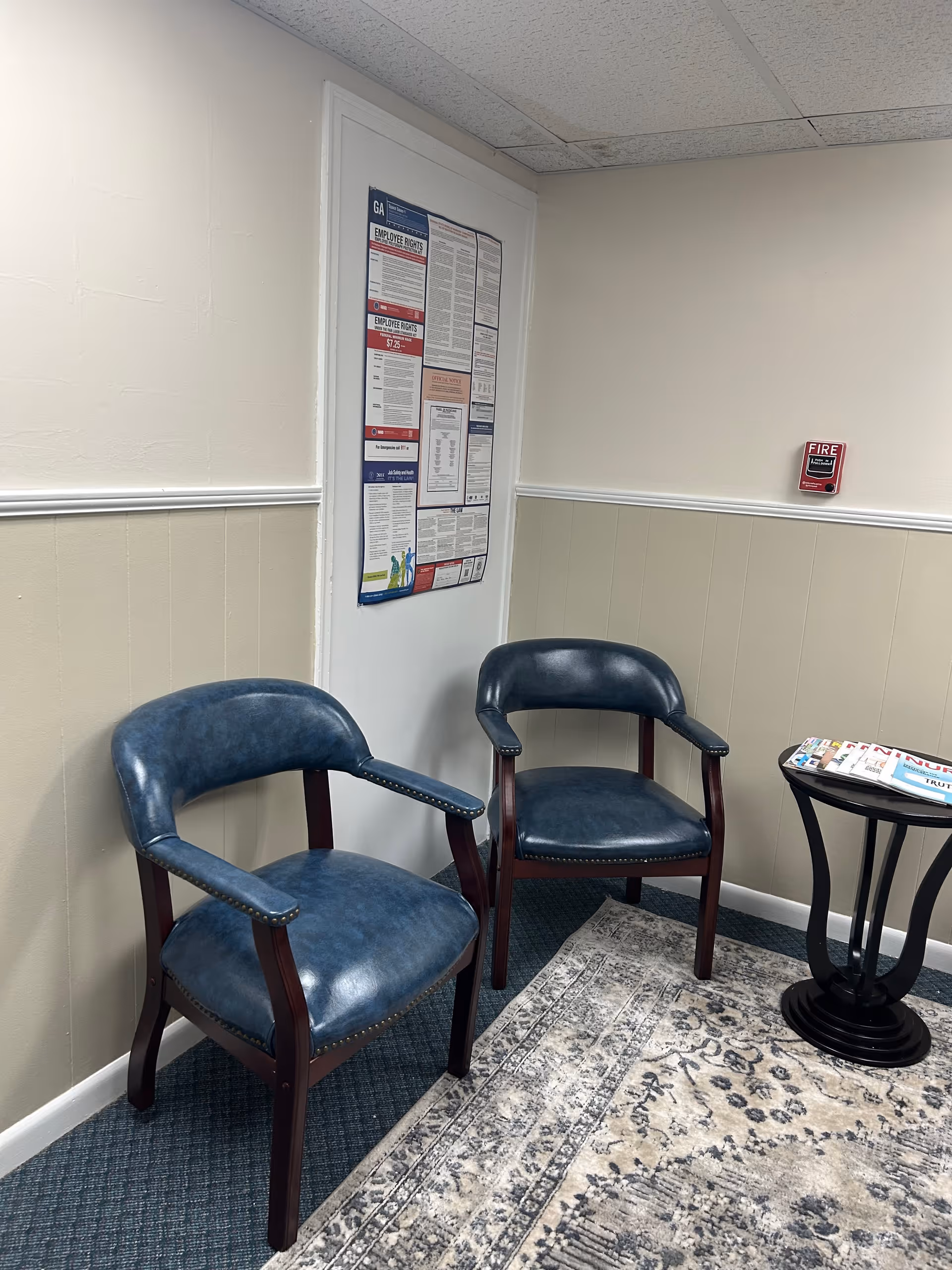Two blue chairs in a small waiting area with a side table and bulletin board.