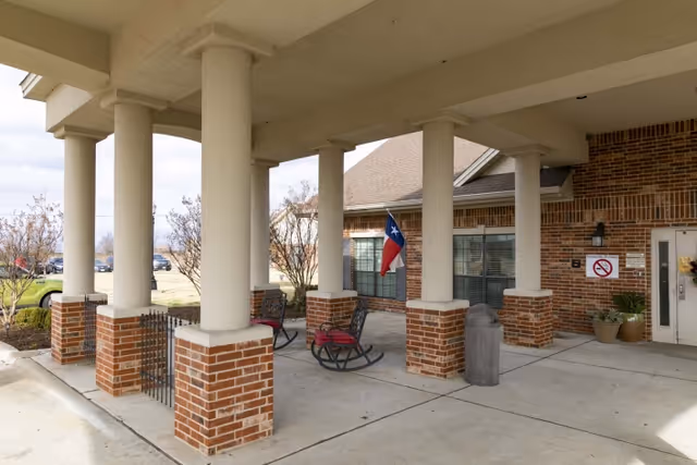 Covered brick entrance with large round columns, rocking chairs, and a Texas flag displayed near the doorway.