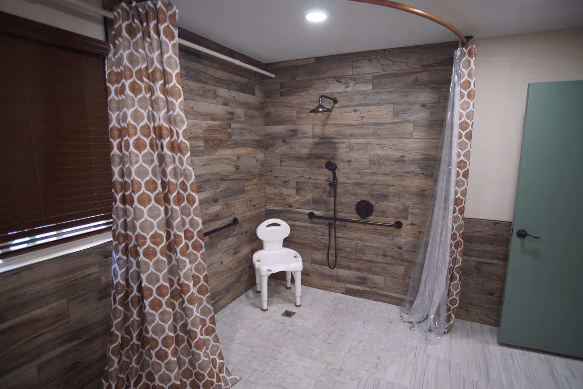 Accessible shower area with wood-textured wall panels, a white shower chair, a handheld showerhead, and grab bars. There is a curved shower curtain rod with a patterned brown and white shower curtain partially drawn. A green door is visible on the right side and a window with closed brown blinds is on the left.