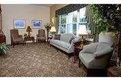 Cozy lounge with sofa, chairs, lamps, and a large windowed wall in a senior facility.