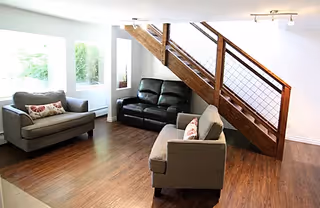 Bright living room with three gray sofas, hardwood floors, and a wooden staircase with cable railing under a large window.