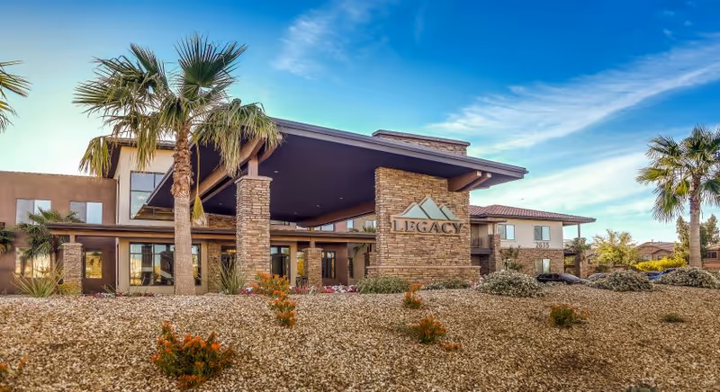Modern senior living facility exterior with stone columns, palm trees, and an entrance sign.