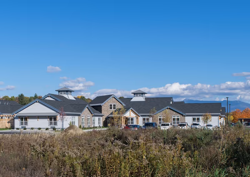 Exterior view of a single-story senior living facility building with multiple peaked roofs and cupolas, surrounded by a parking lot with several cars and natural vegetation in the foreground under a clear blue sky.