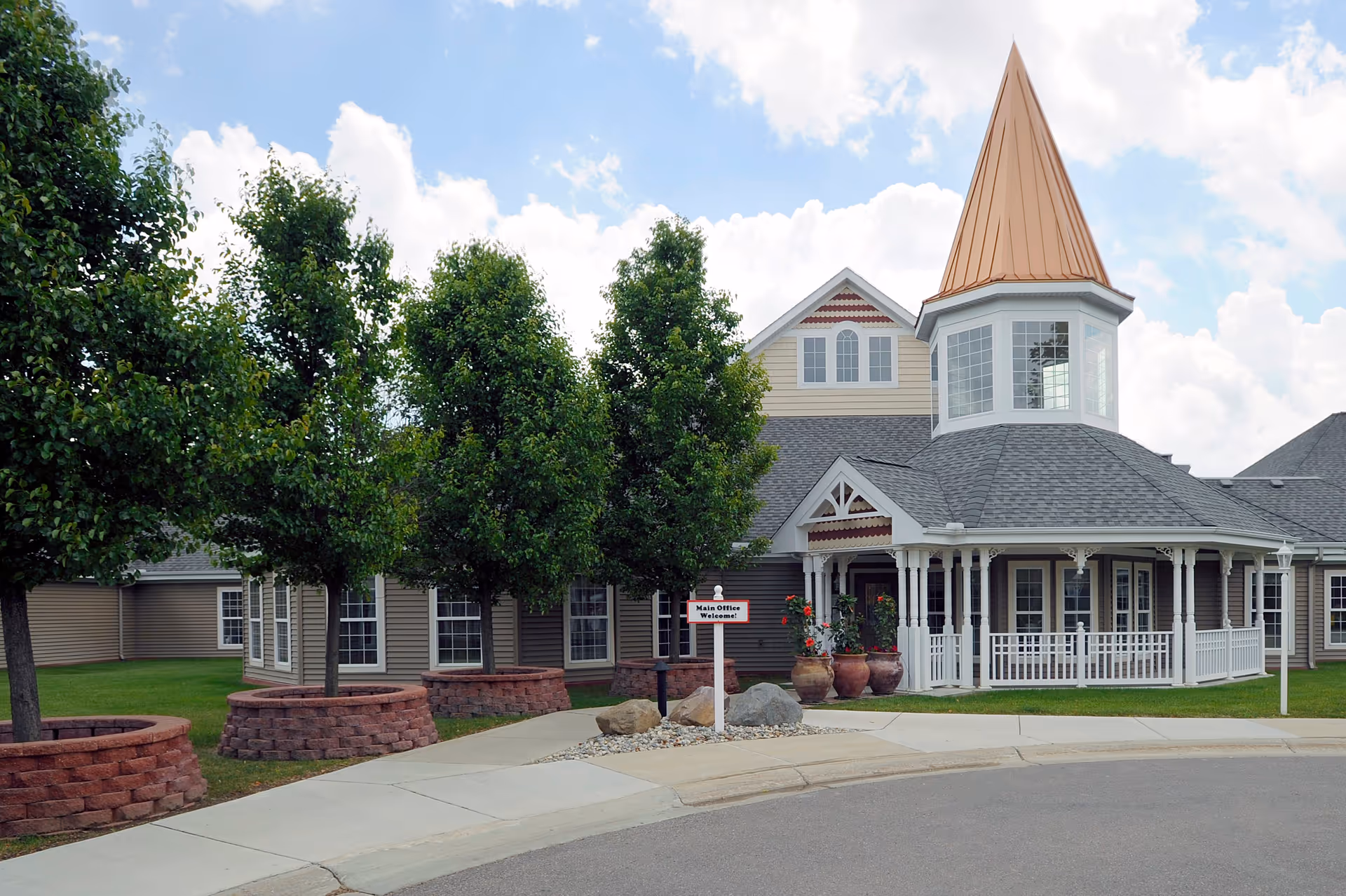Exterior view of a senior living facility building with a pointed turret roof, multiple windows, a white porch railing, and several trees planted in circular brick planters along the sidewalk. A small sign near the entrance reads 'Main Office Welcome!'.